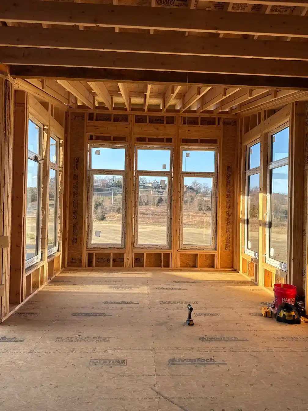 Interior room under construction with exposed wood framing, floor-to-ceiling windows, and open views across the building site