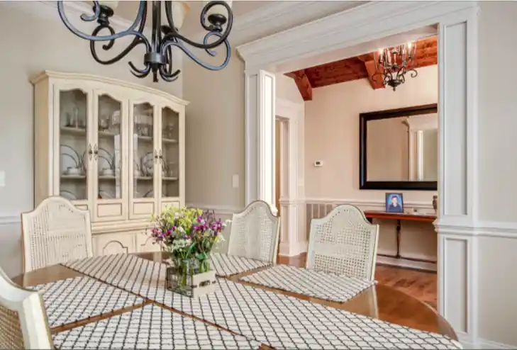 Traditional dining room with glass-front china cabinet, chandelier, paneled trim, and an adjacent room with wood ceiling beams