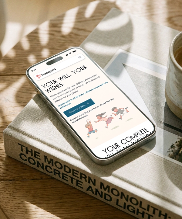 Smartphone displaying a website titled 'YOUR WILL. YOUR WISHES.' on a book titled 'THE MODERN MONOLITH: CONCRETE AND LIGHT'. The site offers legal will creation, showing a 'Start Your Will' button and cartoon illustrations of happy children running. The scene includes natural light with shadows and a cup on the side.