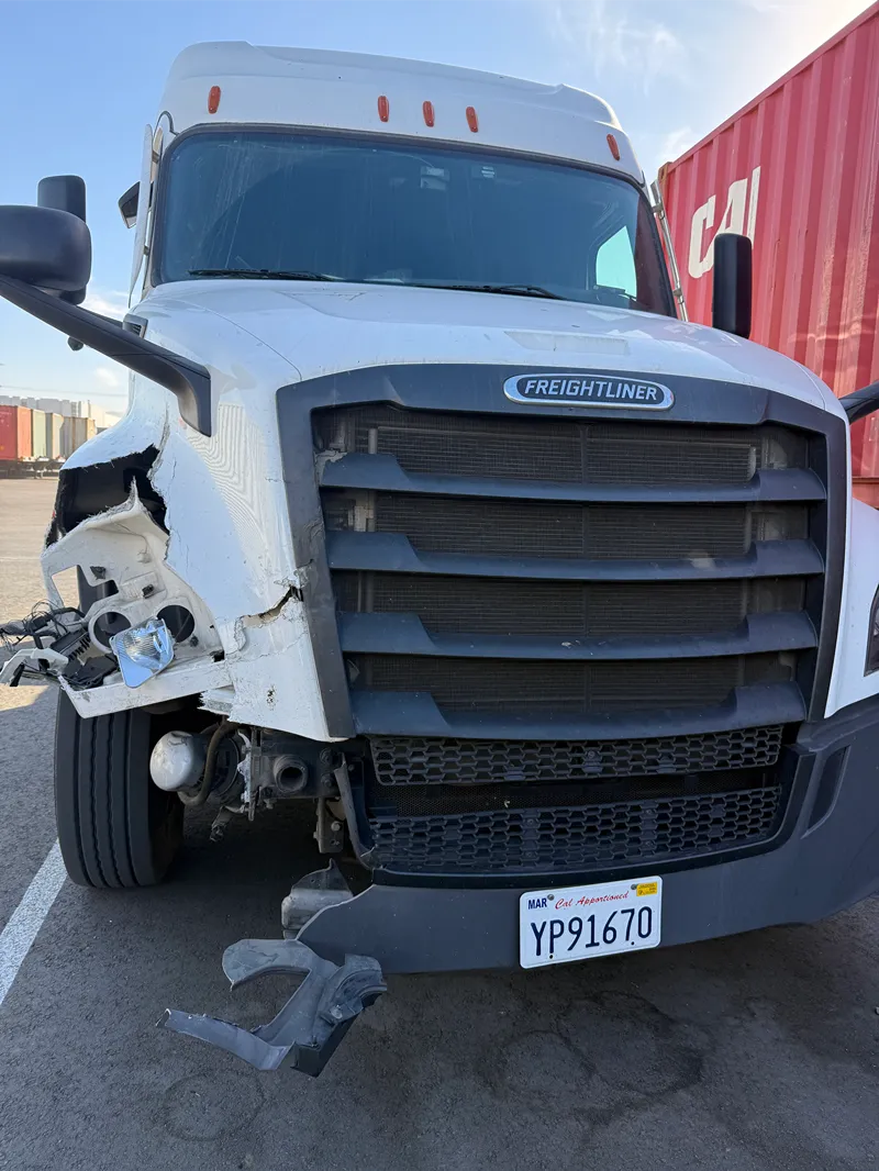 Yard collision damage to a Freightliner truck’s front left corner, with broken panels and exposed headlight components.