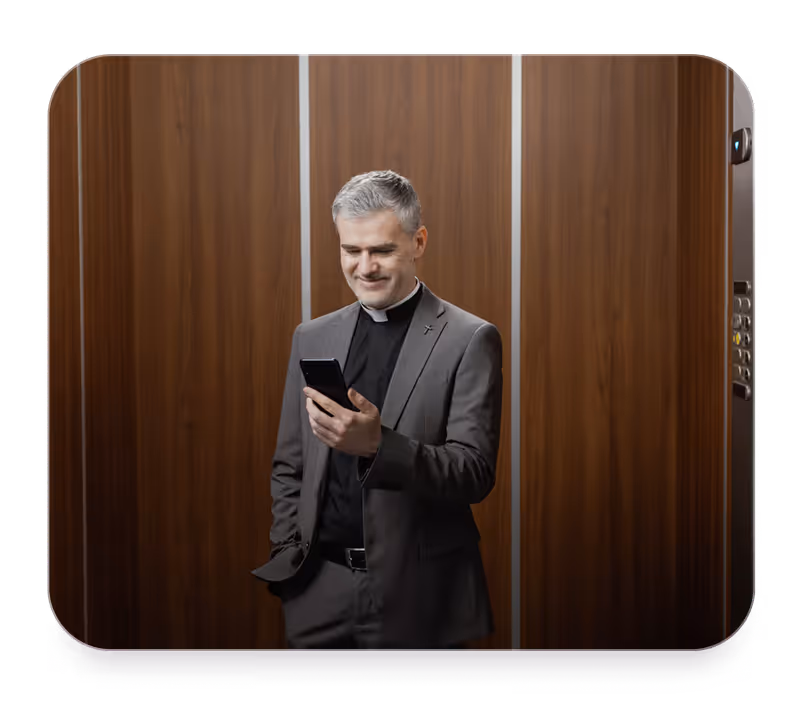 Smiling priest in a gray suit looking at his smartphone inside a wood-paneled elevator.