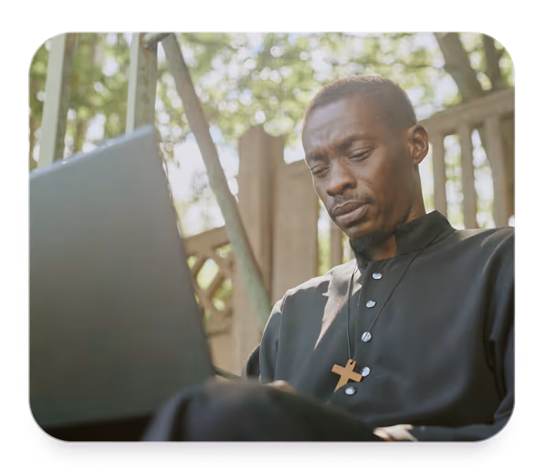 Man wearing a black clerical shirt with a wooden cross necklace using a laptop outdoors.