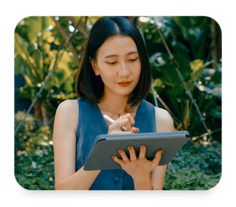 Woman with shoulder-length hair uses a stylus on a tablet outdoors surrounded by green plants.