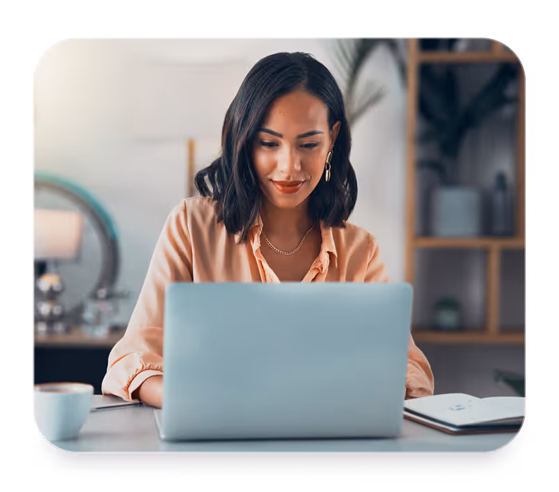 Woman with dark hair using a laptop at a desk with a notebook and coffee cup nearby.