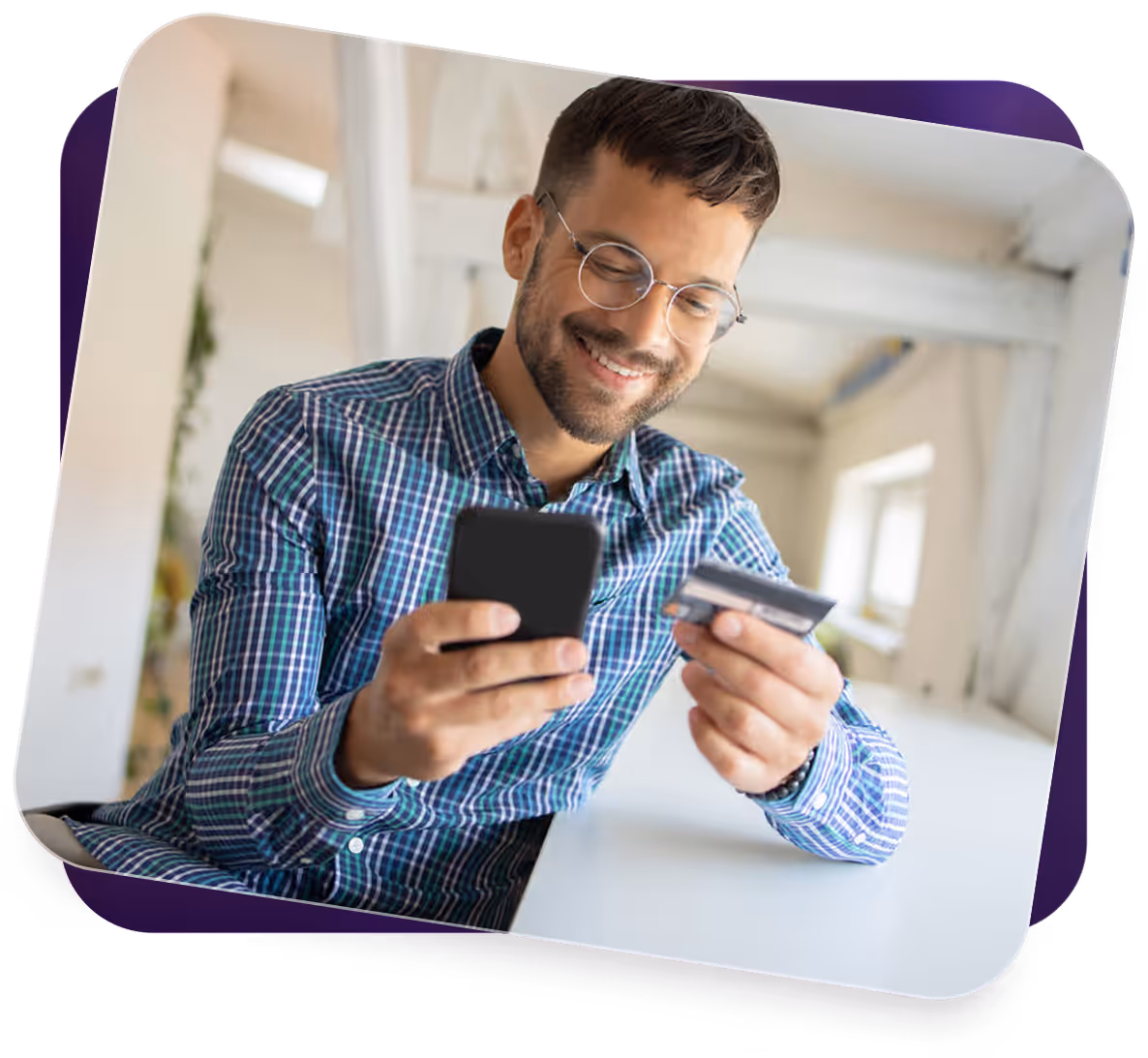 Smiling man with glasses holding a phone and credit card, sitting at a white desk in bright room.