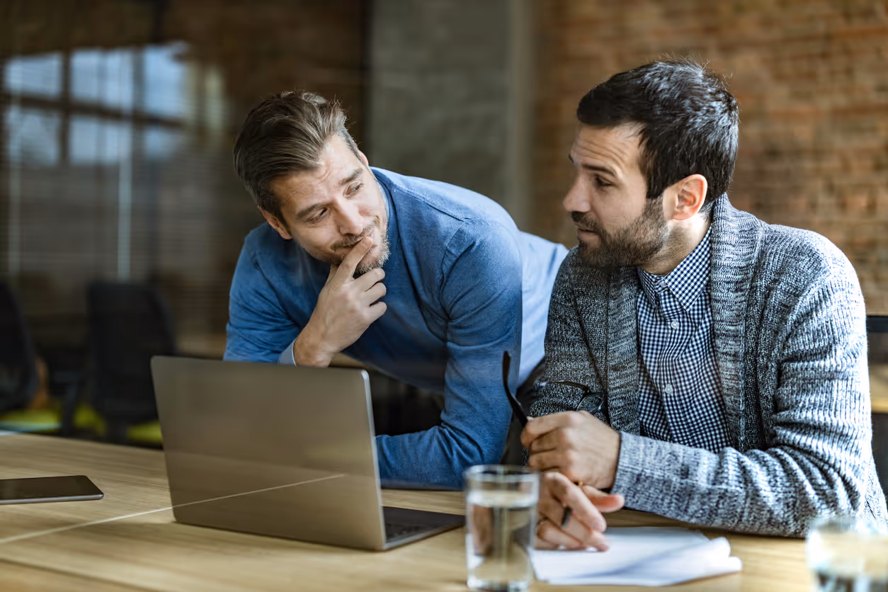 Two men discussing while looking at a laptop on a wooden desk in a modern office.