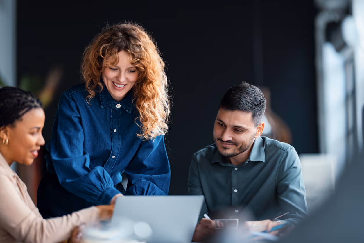 Three colleagues smiling and discussing something on a laptop in a modern office setting.