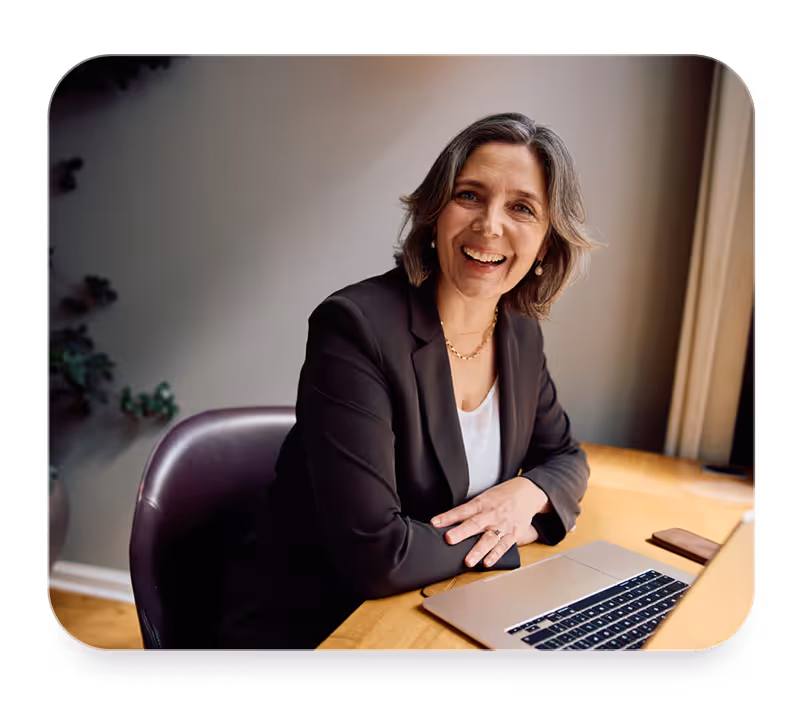 Smiling woman with gray hair in business attire sitting at a desk with an open laptop.