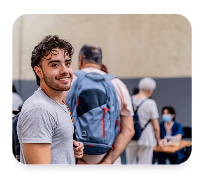 Young man smiling at camera with backpack in line, people and volunteer wearing mask in background.