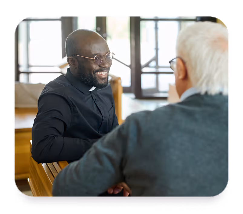Smiling Black priest wearing glasses and clerical collar talks to an older white man in a gray sweater.