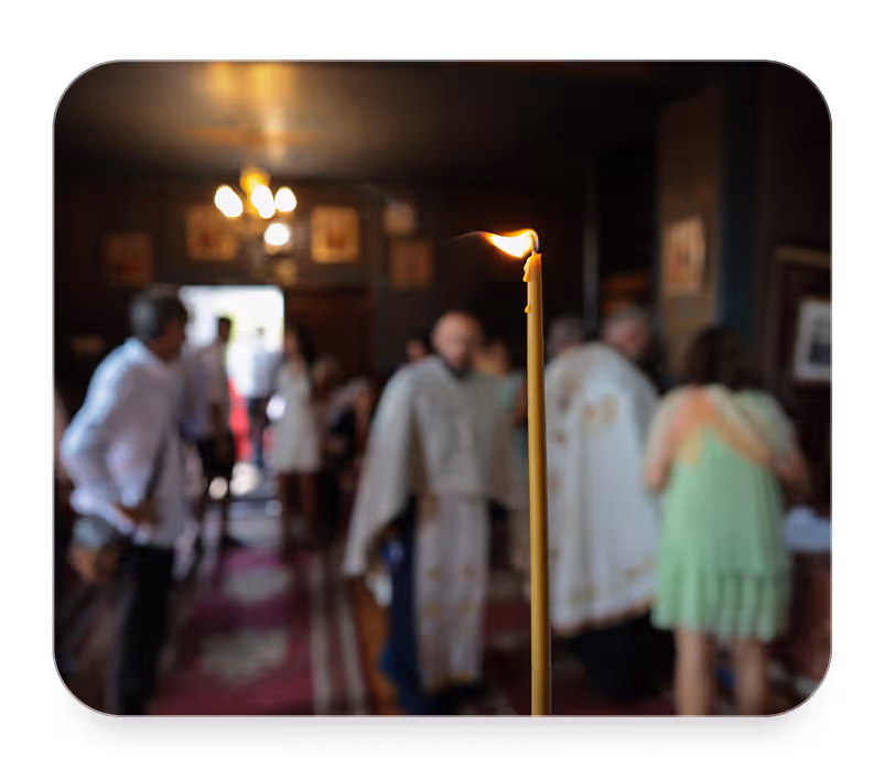Lit candle in focus inside a dimly lit church with blurred people and clergy in the background.