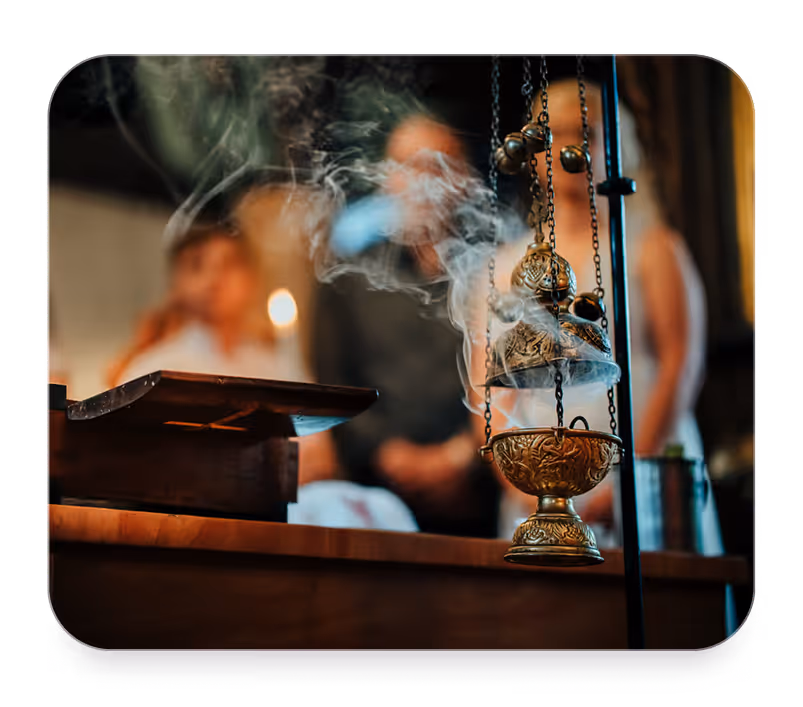 Smoking ornate gold incense burner hanging in the foreground with blurred people and candle in background.