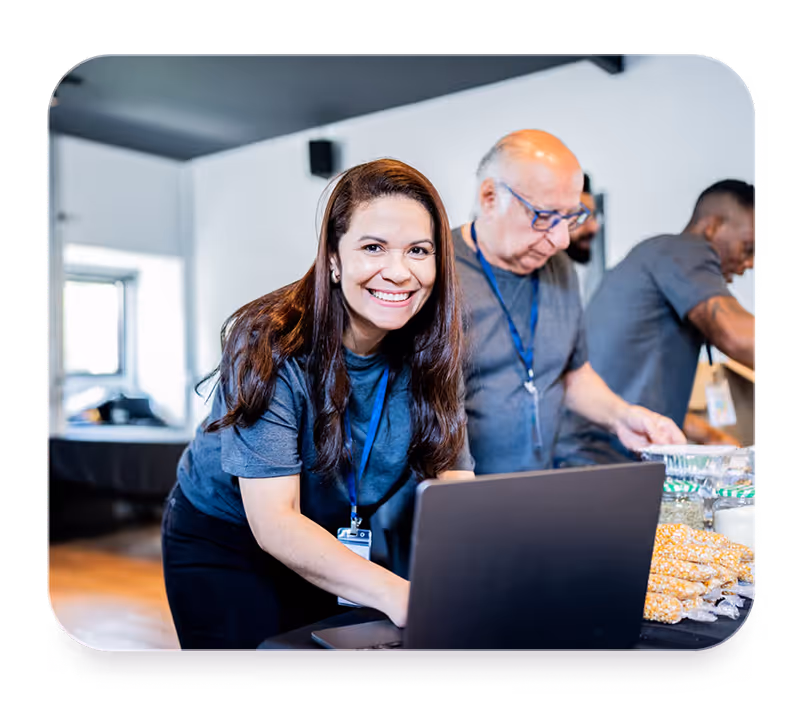 Smiling woman volunteering with others organizing supplies on a table with a laptop indoors.