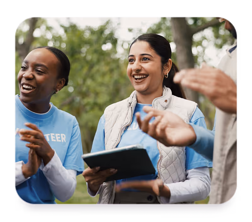 Smiling volunteers outdoors, one holding a tablet, wearing blue shirts and a white vest.
