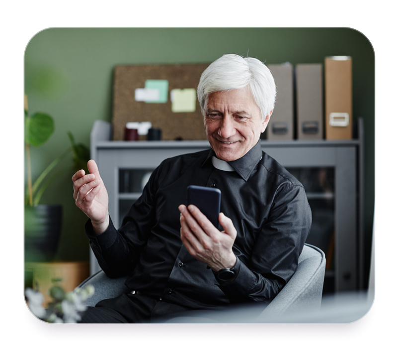 Smiling older man in priest collar sitting in a chair looking at his smartphone in an office.