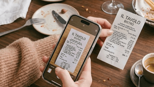 Person photographing a restaurant receipt with the KleerCard app on their smartphone on a wooden table with dishes and coffee cup.