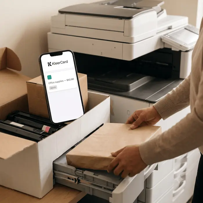 Person handling a package near a printer and phone showing KleerCard expense of $42.80 for office supplies.