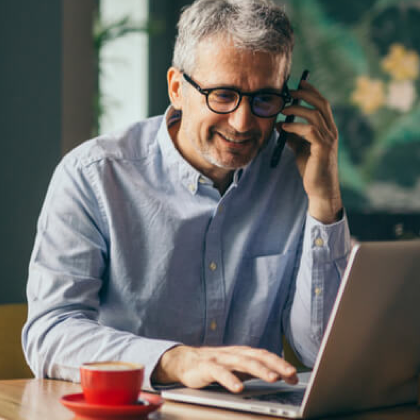 Smiling man wearing glasses talks on phone while using laptop at a table with a cup of coffee.