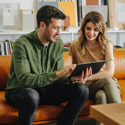 Man and woman sitting on a couch looking at and discussing a tablet screen together.