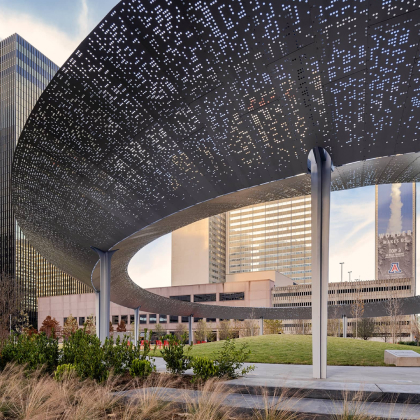 Curved metal canopy with perforations over urban park in front of modern glass and concrete buildings.