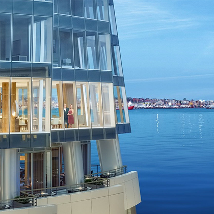Modern glass building over water with two people visible inside near a coastal cityscape.