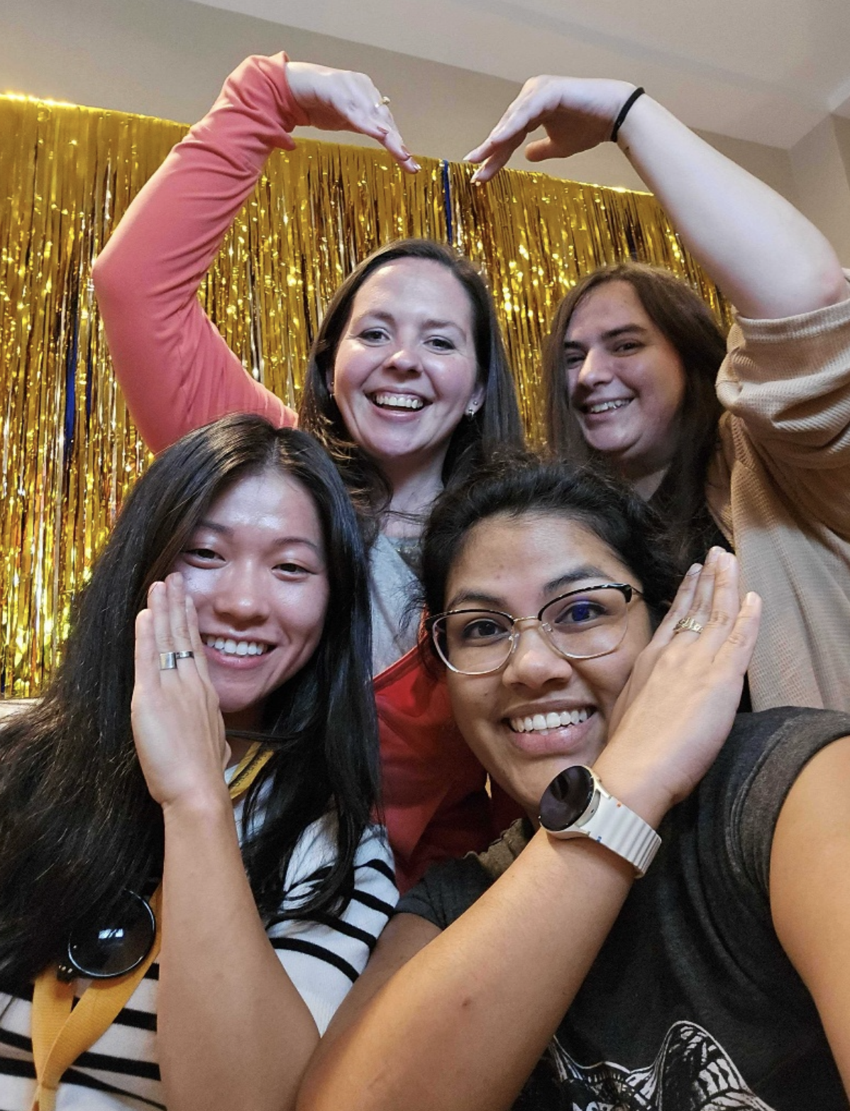 Four smiling women posing cheerfully in front of golden tinsel backdrop making heart shapes with their hands.