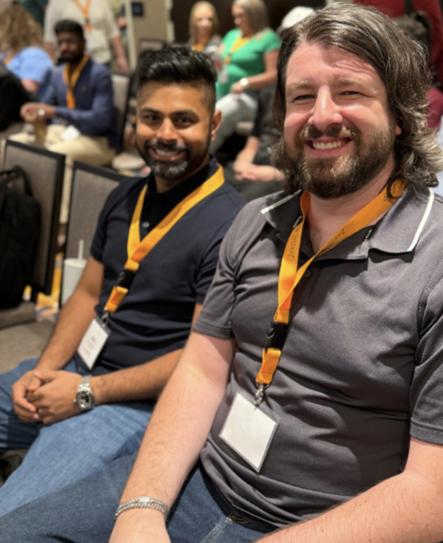 Two men smiling and wearing orange lanyards with badges sitting in a conference room.