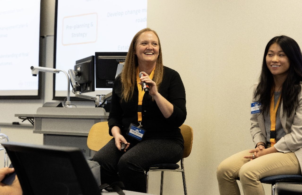 Two women with lanyards sit on chairs, one speaking into a microphone in a presentation room.