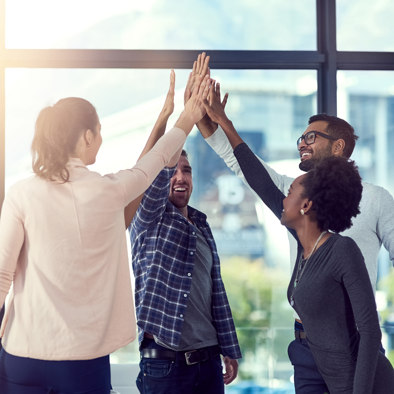 Four diverse young adults in casual clothing standing in a bright office space raising their hands together in a group high-five, smiling and showing teamwork.