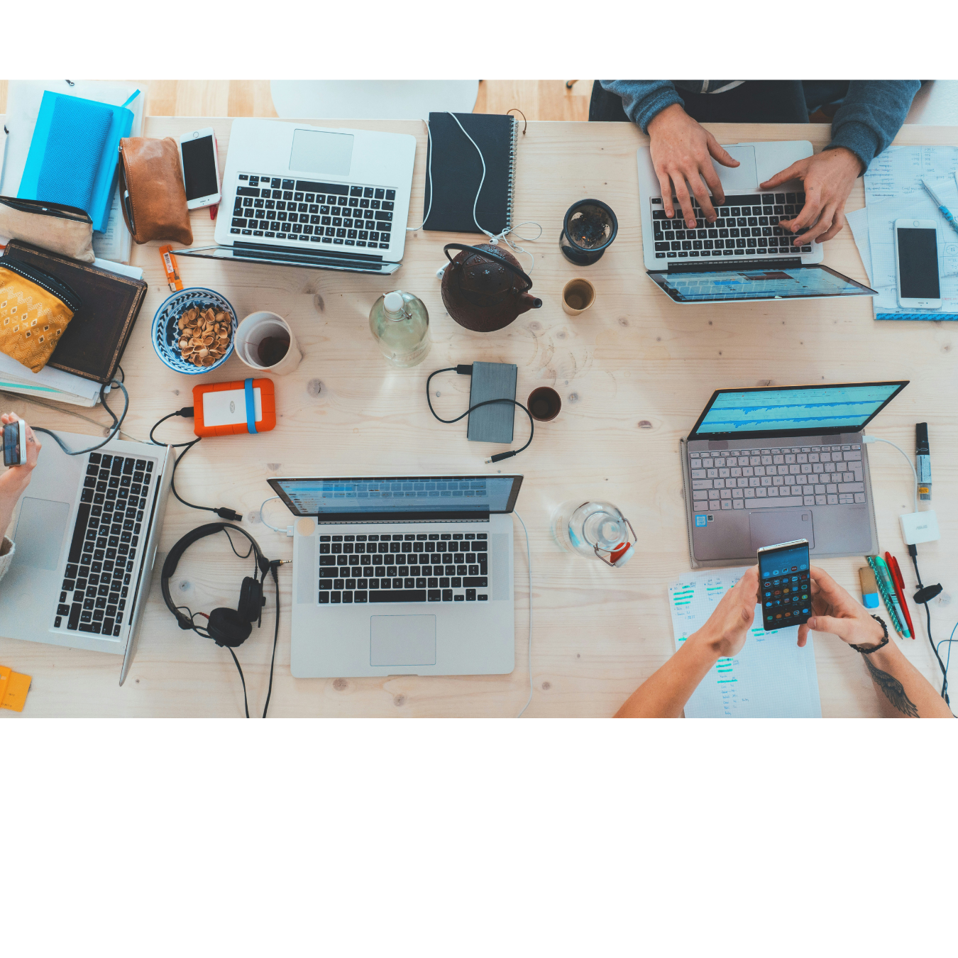 Top view of a wooden table with four laptops, headphones, smartphones, notebooks, pens, a teapot, cups, and cords, with people working collaboratively.