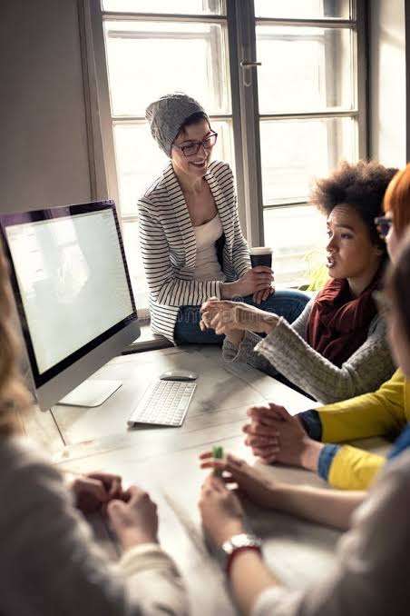 Group of young adults in a bright room around a table with a computer, engaged in discussion; one woman wearing glasses and a beanie sits on the windowsill holding a cup and smiling.