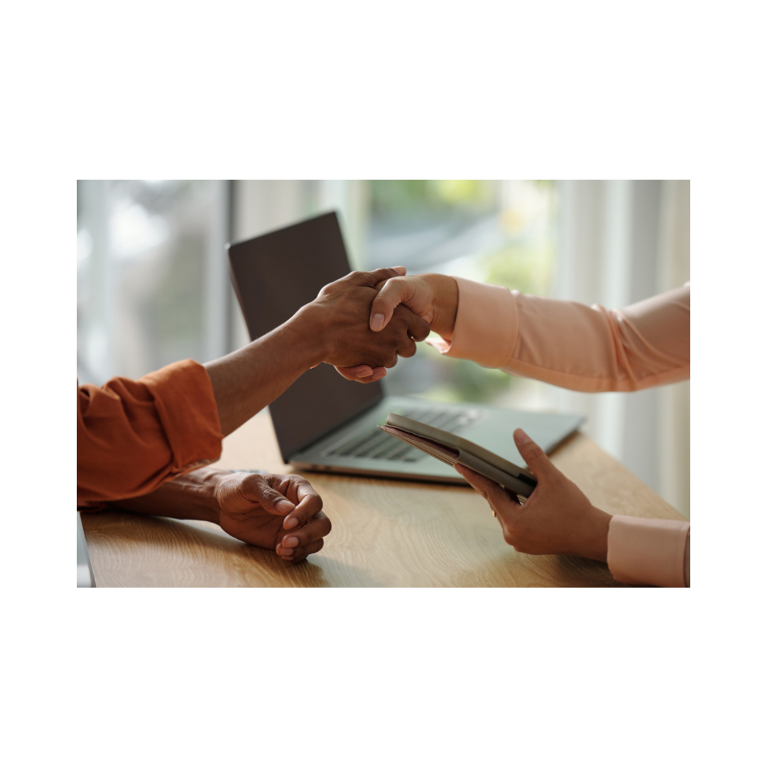 Two people shaking hands over a wooden table with a laptop in the background, one person holding a tablet.