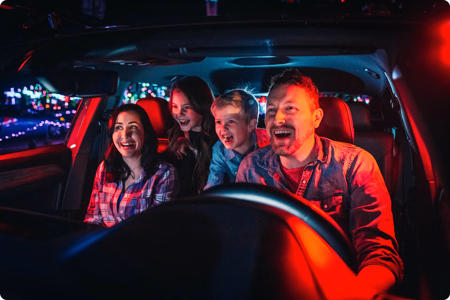 Smiling family of four in a car at night with colorful light reflections outside.