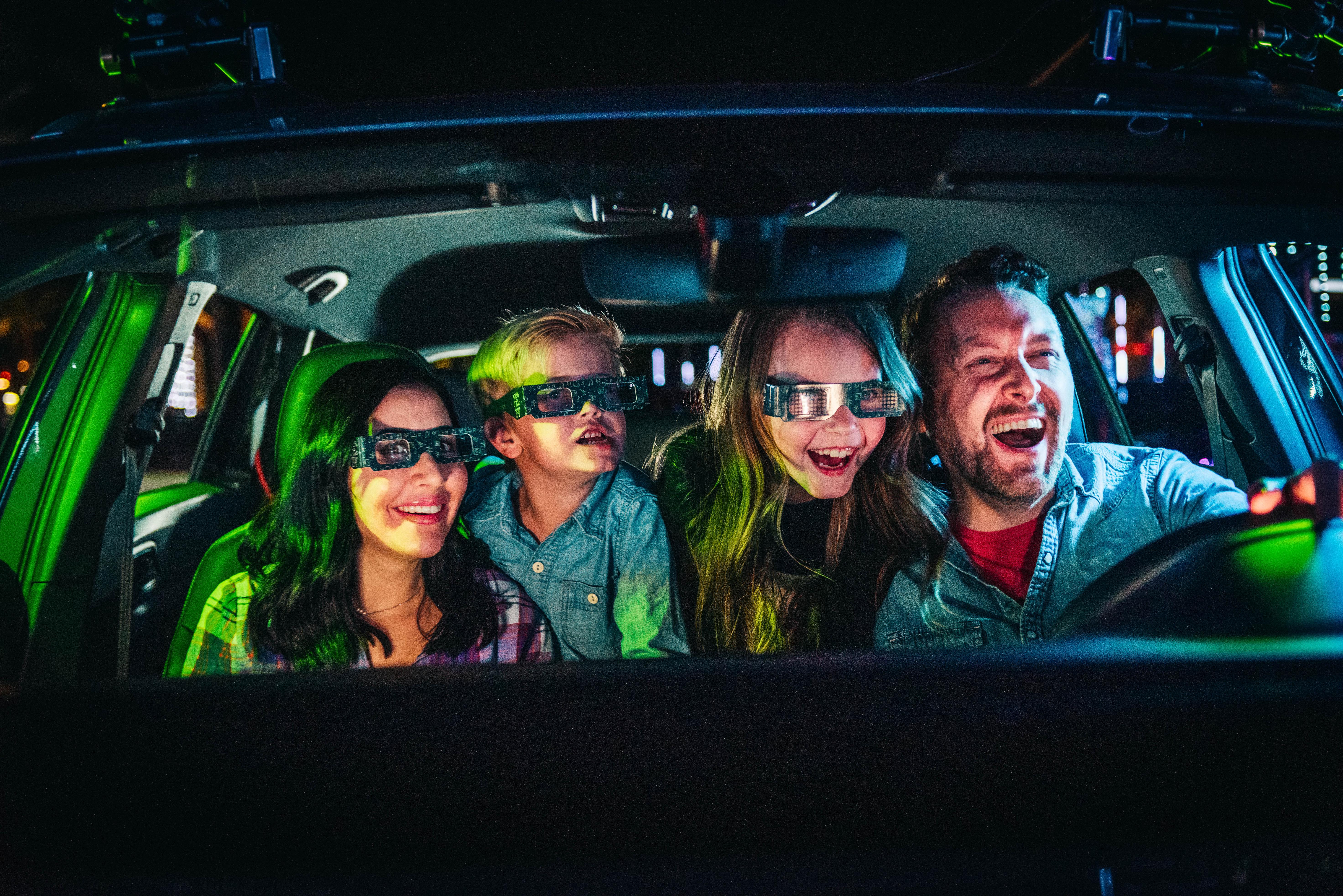 Smiling family of four in a car at night with colorful light reflections outside.