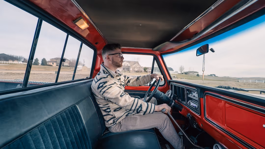 Man wearing glasses and a patterned sweater driving a red pickup truck with a rural landscape outside.