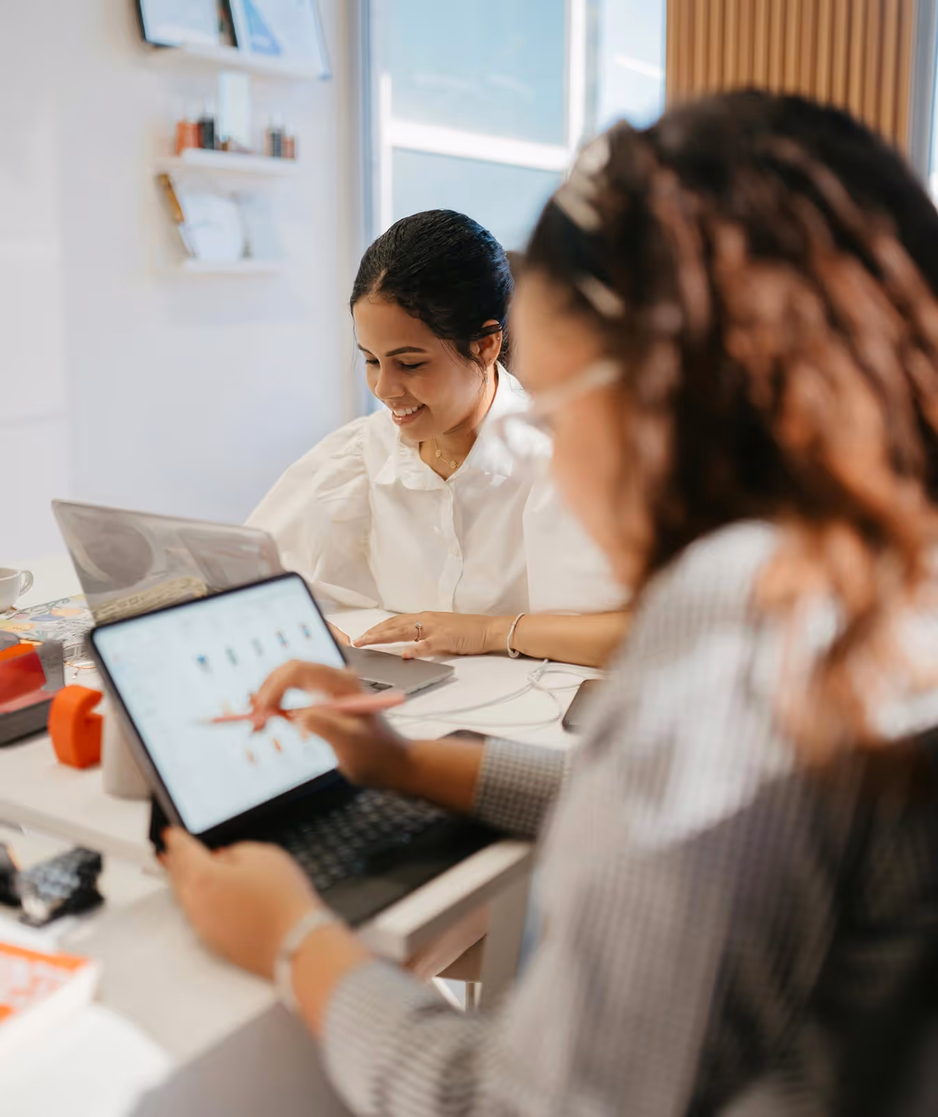 Two women working on laptops at a desk, one smiling while the other uses a digital pen on a tablet.