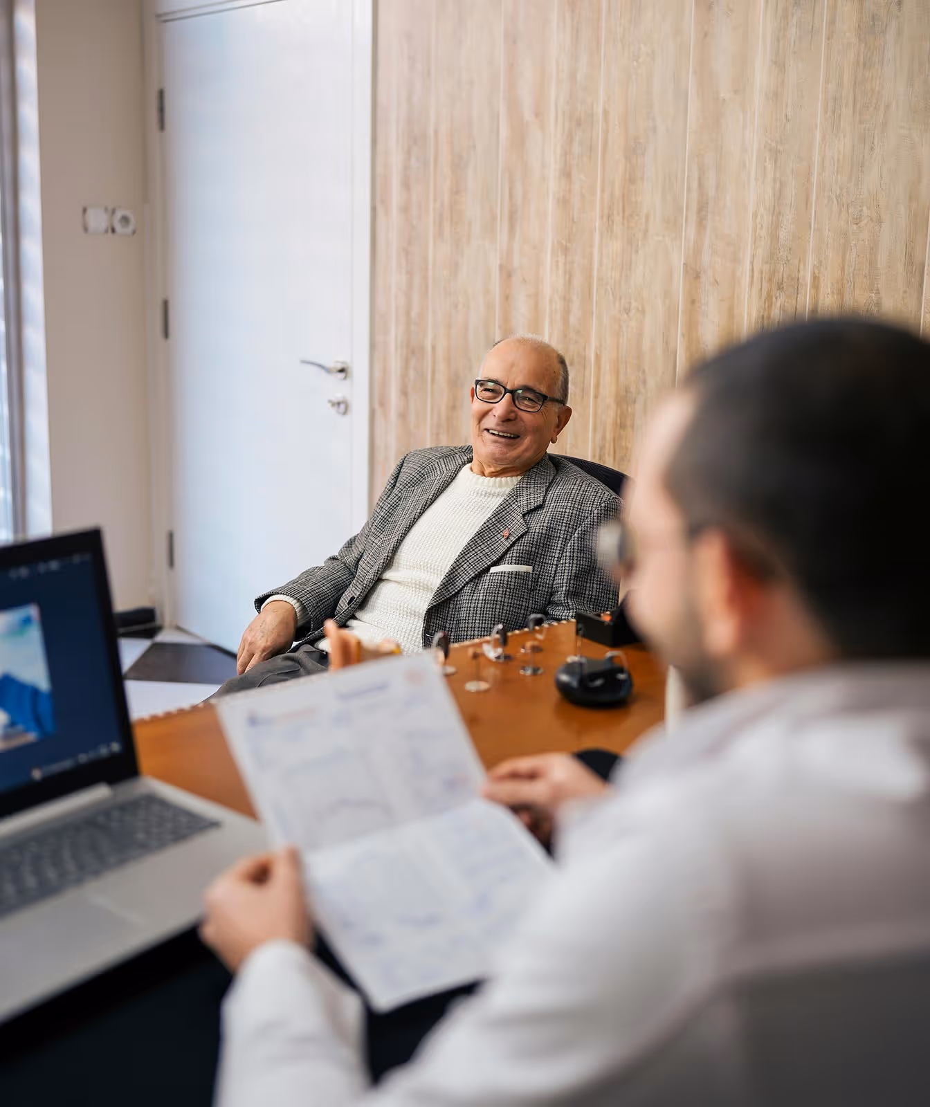 Older man smiling during consultation with a professional holding a document in an office setting.