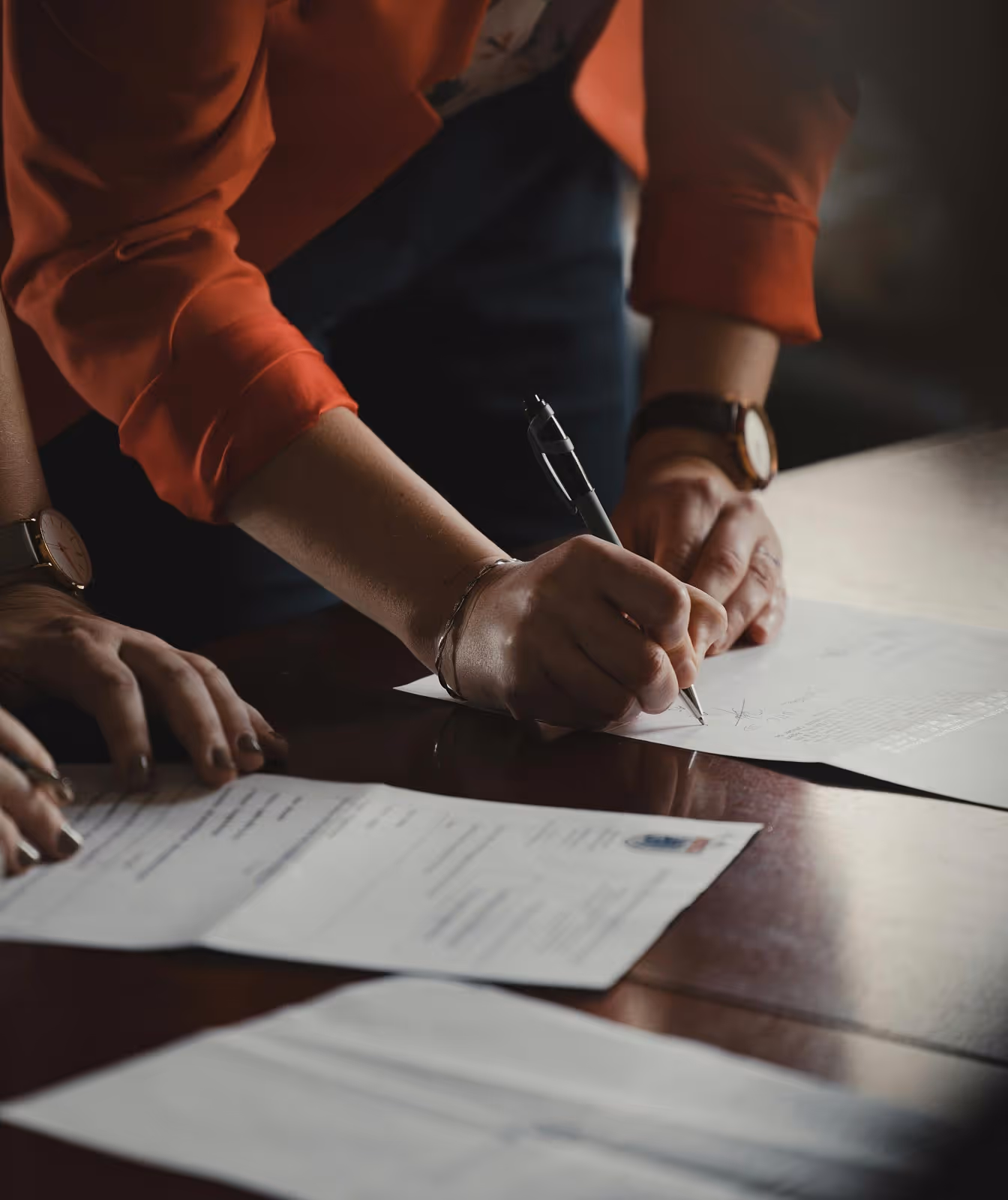 Person wearing a red sleeve signing documents on a wooden table with another person nearby.