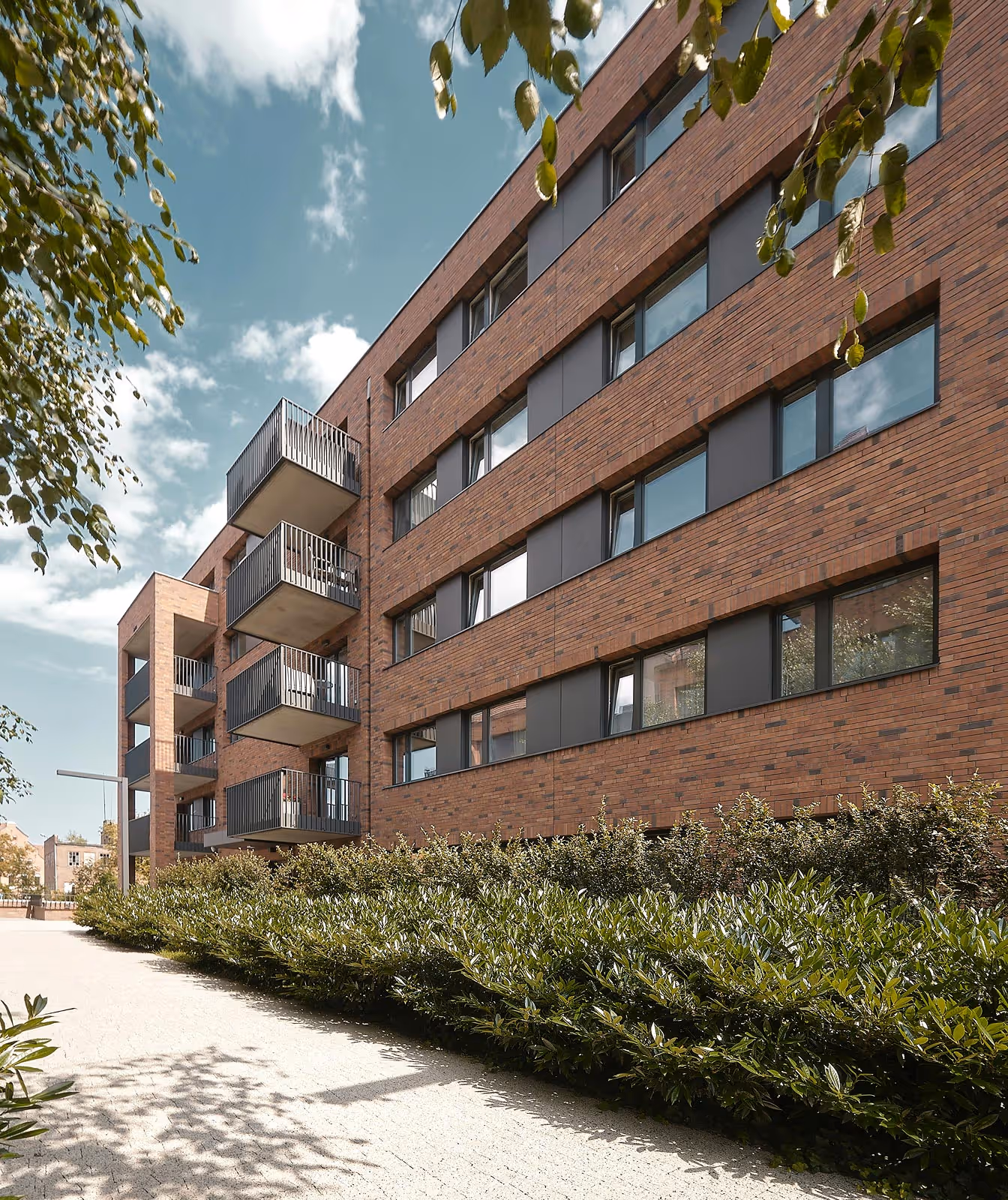 Modern four-story brick apartment building with balconies and bushes along the sidewalk.