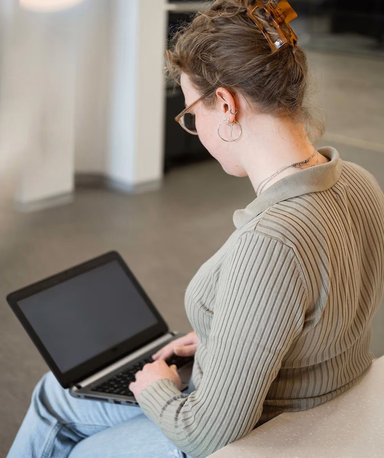 Woman with glasses and hair clip typing on a laptop while seated indoors.