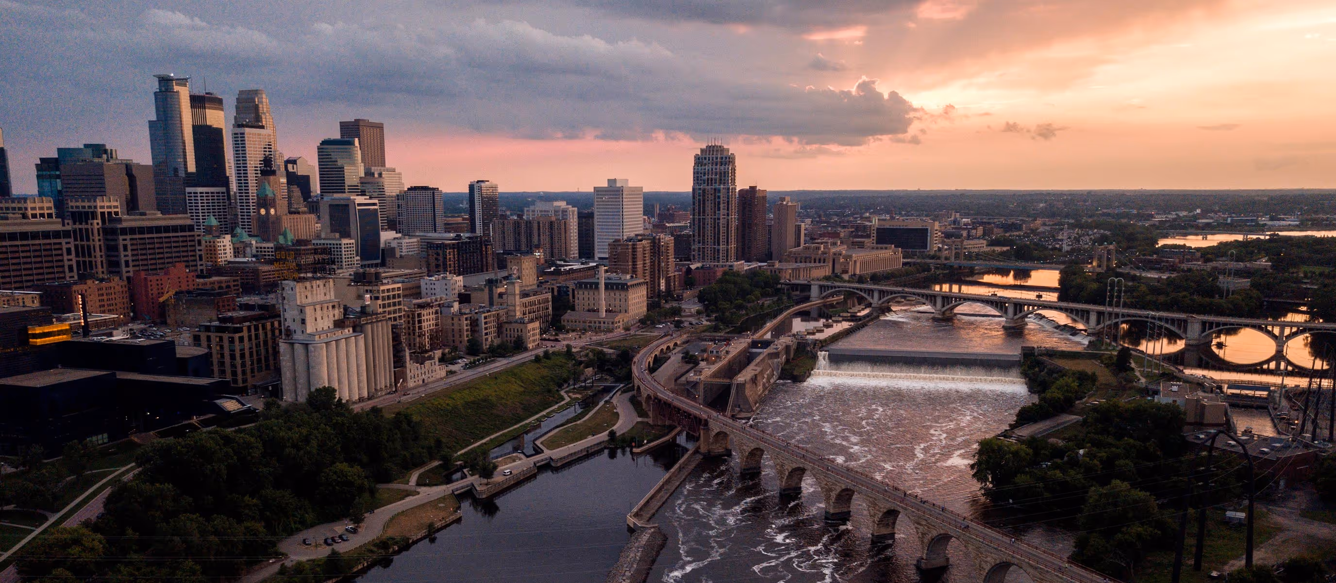Minneapolis city skyline at sunset with bridges over the Mississippi River and urban buildings.