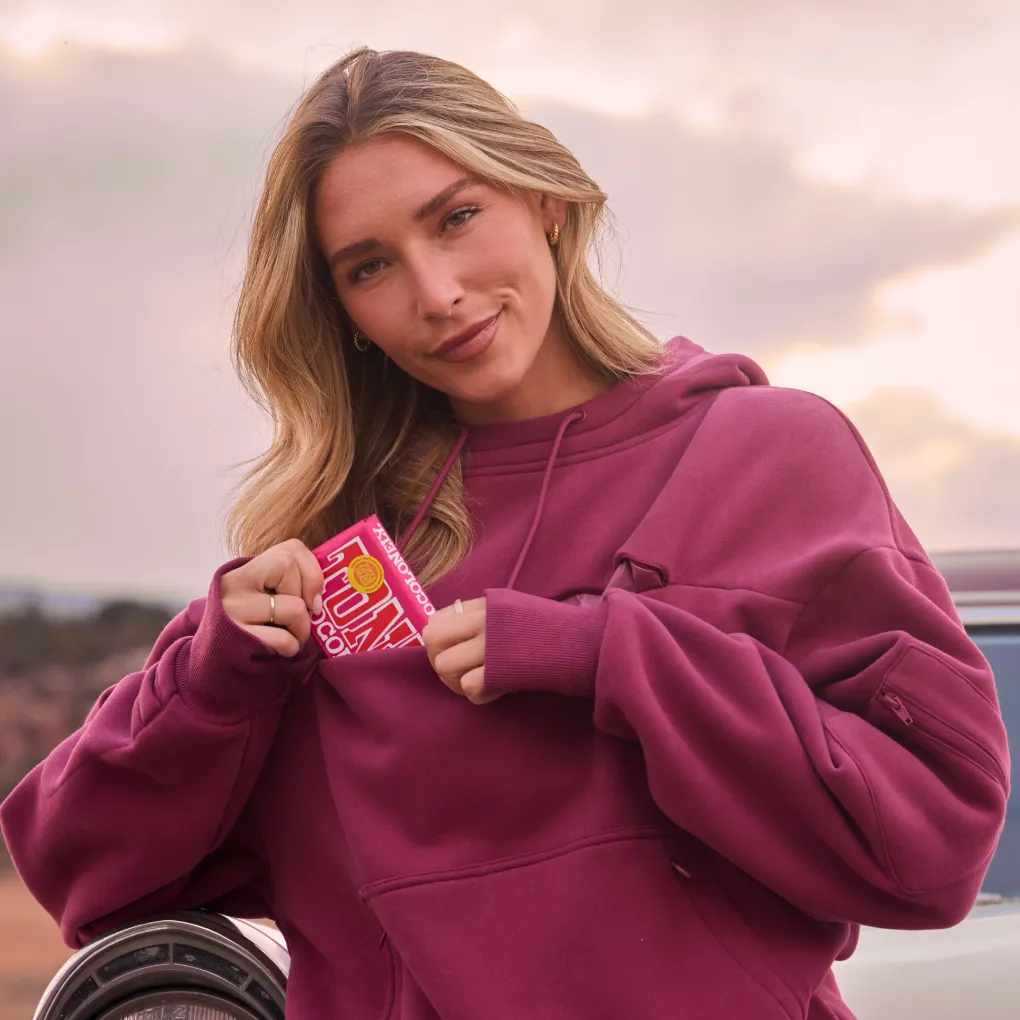Woman slipping a chocolate bar into the zippered food-safe chest pocket of a berry-red Snak Hoodie