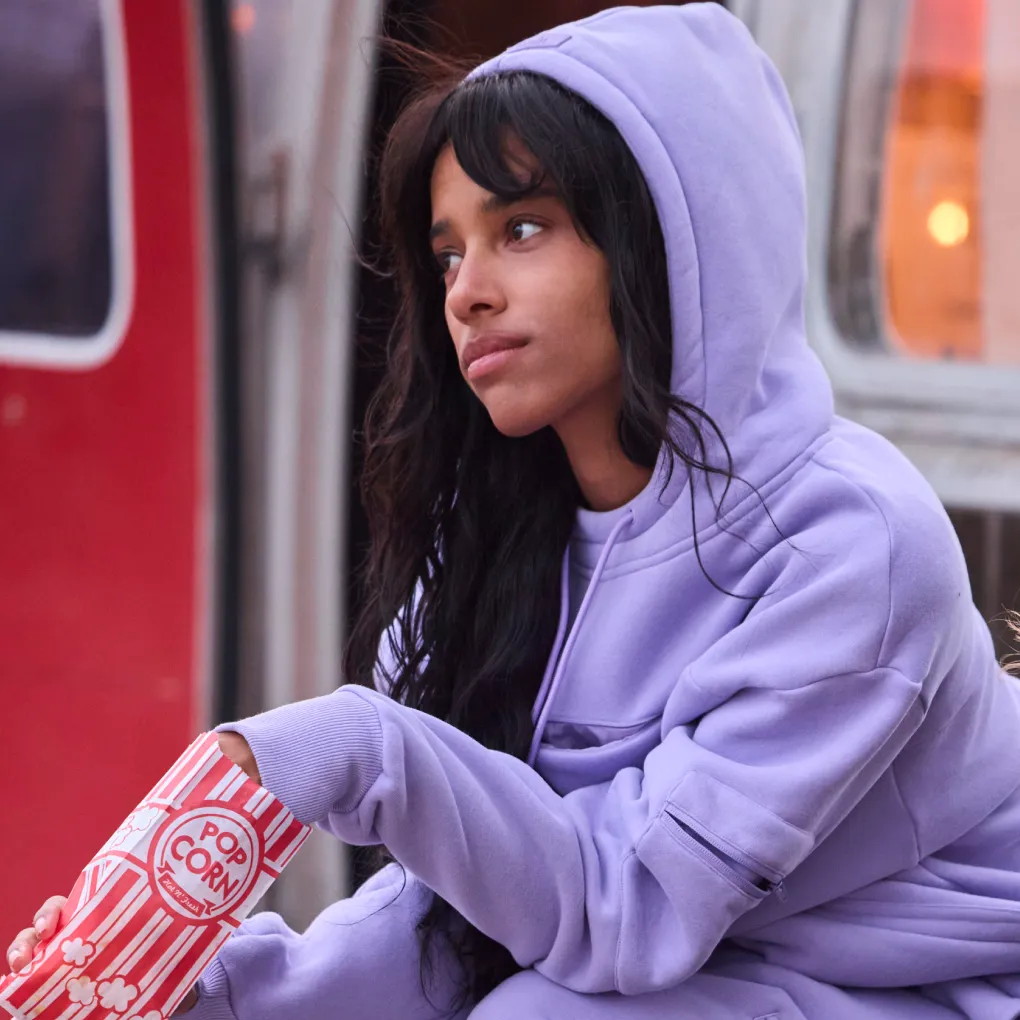 Woman relaxing with the oversized hood of a lavender Snak Hoodie pulled up, holding a striped carton of popcorn