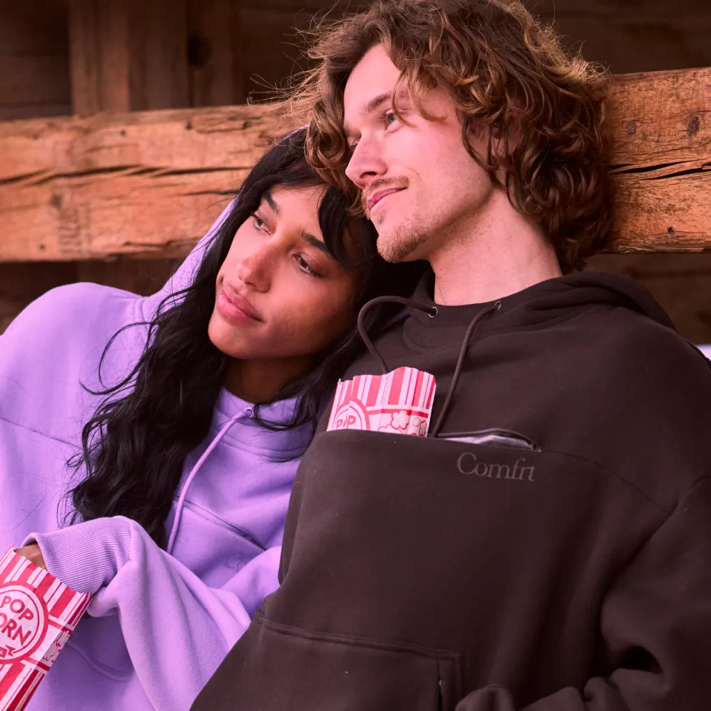 Couple lounging together, a popcorn carton tucked into the chest pocket of his dark brown Snak Hoodie