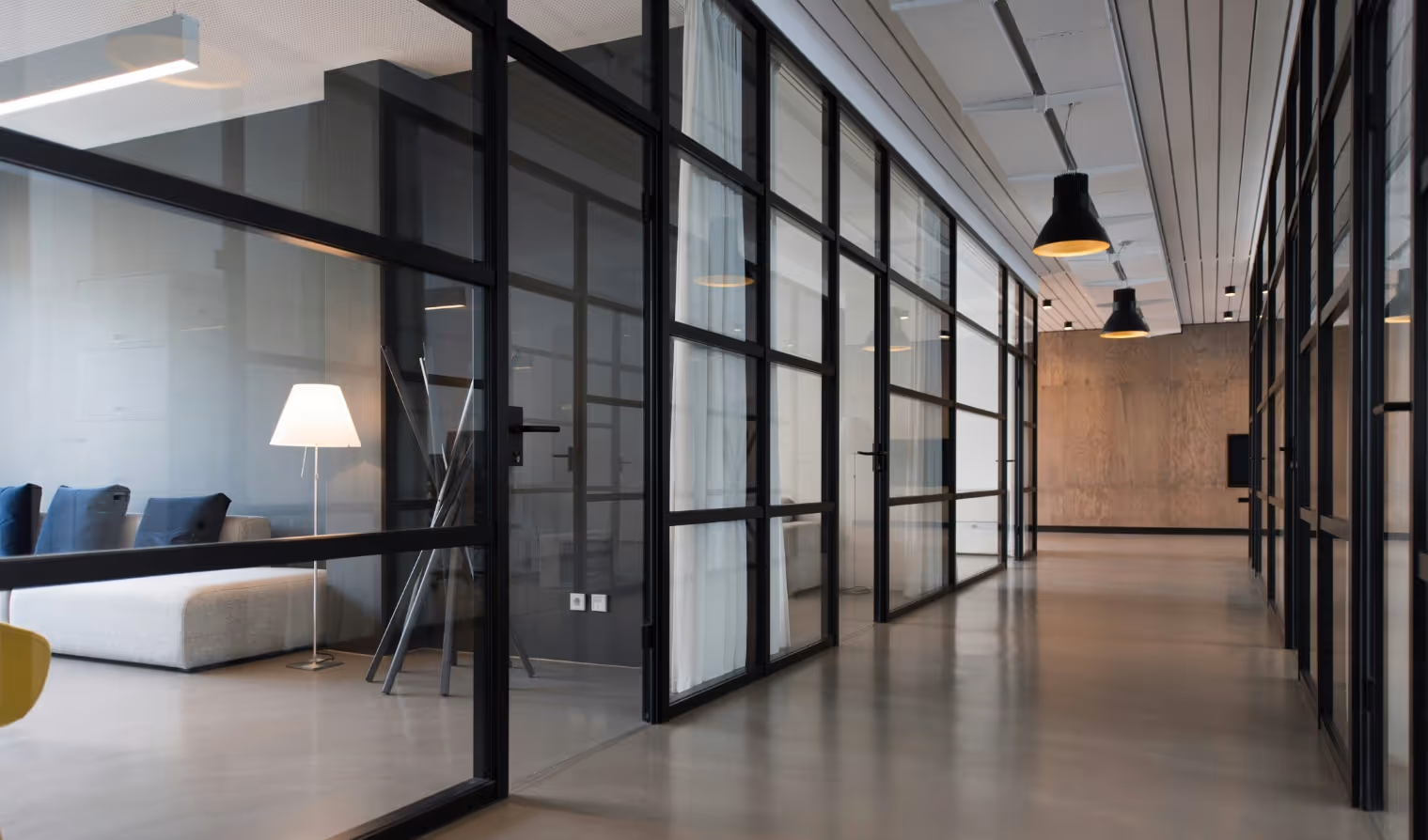 Modern office corridor with glass walls, concrete floor, pendant lights, and visible lounge area with sofa and lamp.