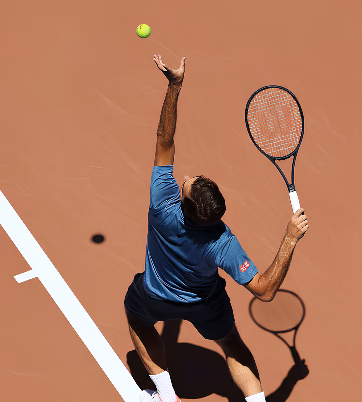 Tennis player in blue shirt tossing ball up for a serve on a clay court from overhead view.