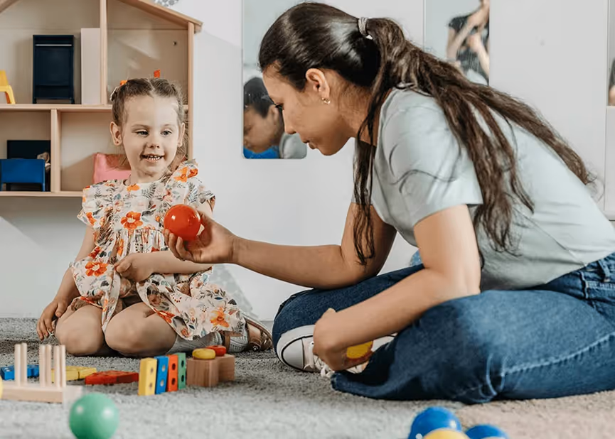 Woman playing with a young girl on the floor surrounded by colorful toys and balls in a playroom.