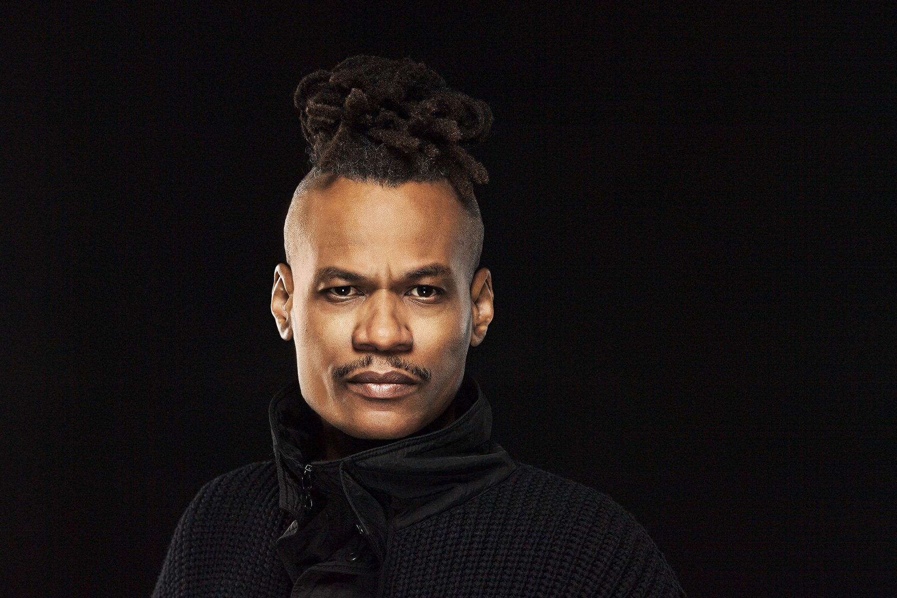The Mighty Cobra in a black knit sweater, locs tied up, studio portrait against black.