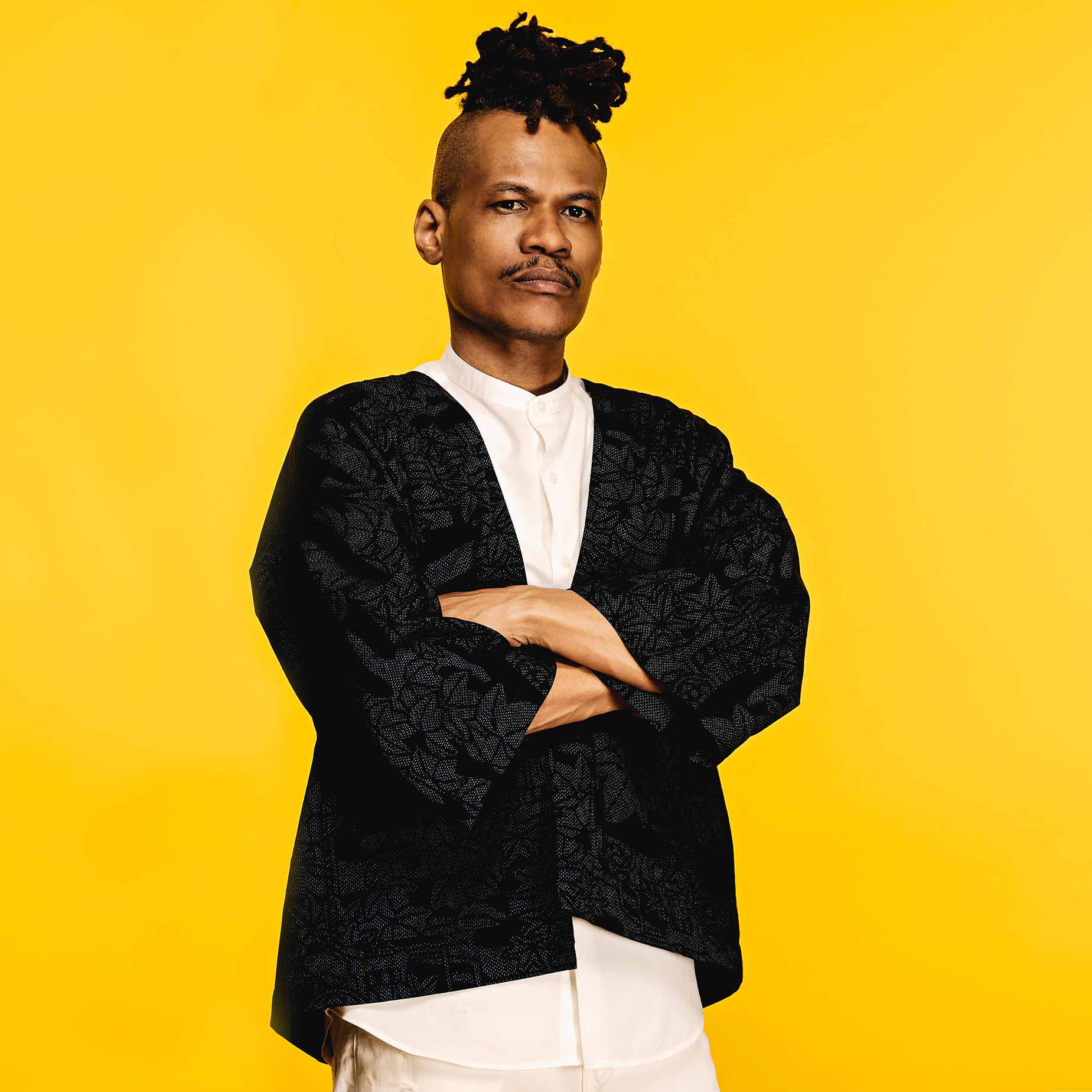 The Mighty Cobra with arms folded, in a black patterned jacket over white, studio portrait on a yellow background.