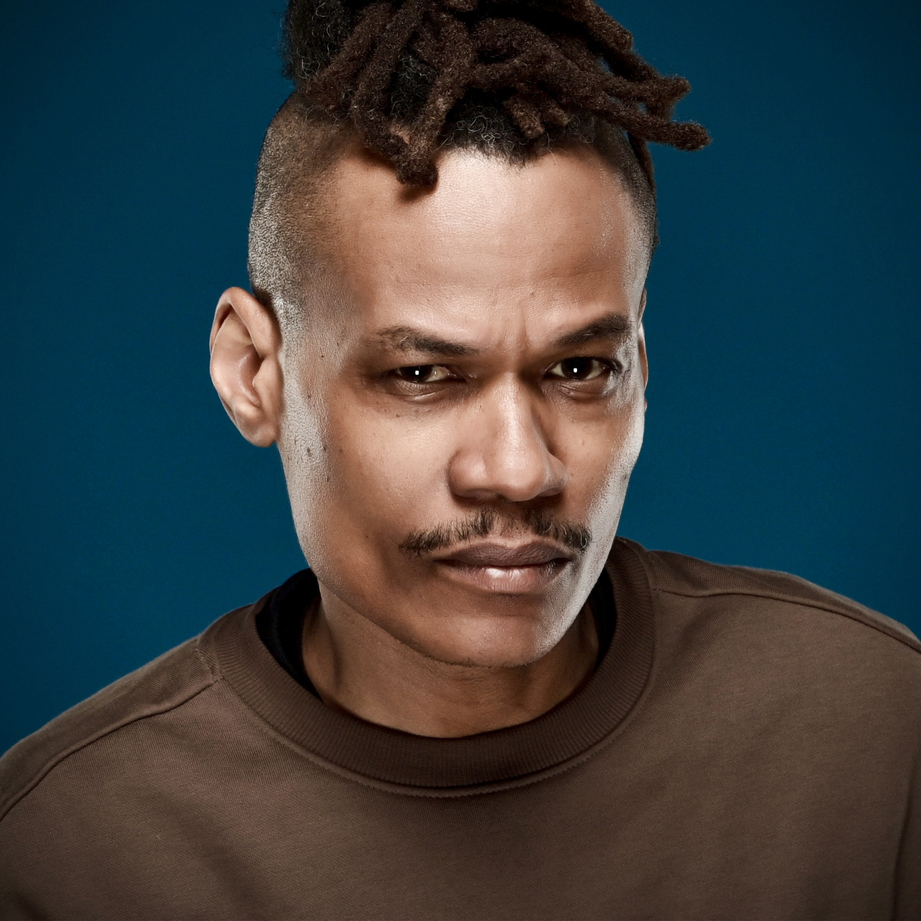 The Mighty Cobra in a brown sweatshirt, locs tied up, studio portrait on a deep blue background.